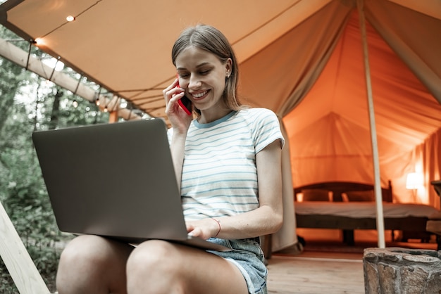 Junge lächelnde blonde Frau, die auf Glamping-Boden mit Laptop sitzt und auf Smartphone spricht. Camping-Lebensstil. Reisen mit geringem Budget. Konzept der Remote-Arbeit. Moderne Internet-Kommunikationstechnologie.