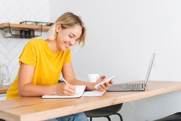 Junge lächelnde blonde Frau im gelben T-Shirt, die zu Hause am Laptop in der Küche arbeitet