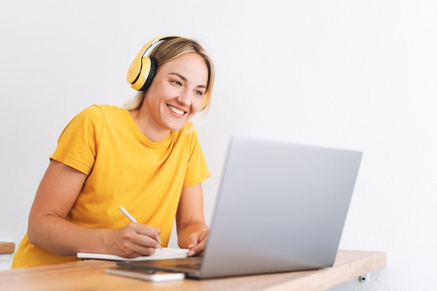 Junge lächelnde blonde Frau im gelben T-Shirt mit Kopfhörern, die zu Hause am Laptop in der Küche arbeitet