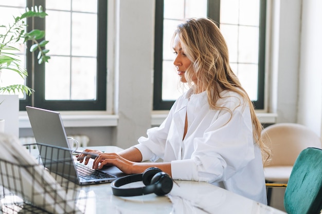 Junge lächelnde blonde Frau mit langen Haaren im stilvollen weißen Hemd, die am Laptop im hellen, modernen Büro arbeitet und über den Videoanruf spricht