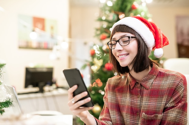 Junge lächelnde brünette Geschäftsfrau in der Weihnachtsmannmütze, die selfie durch Arbeitsplatz mit Weihnachtsbaum macht