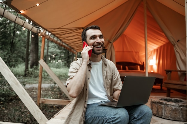 Foto junge lächelnde brünette mann sitzt auf glamping mit laptop sprechen auf smartphone, streaming. camping-lebensstil. reisen mit geringem budget. konzept der remote-arbeit. moderne internet-kommunikationstechnologie.