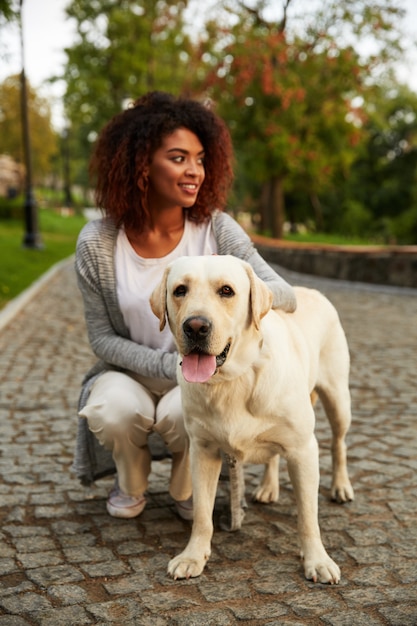 Junge lächelnde Dame in lässiger Kleidung, die Hund im Park sitzt und umarmt