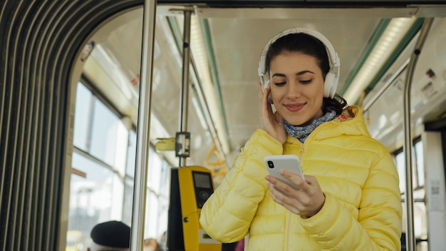 Junge lächelnde Frau bei der Stellung im Zug, in der Tram oder im Bus. Glücklicher weiblicher Passagier, der öffentlich Musik auf einem Transport des Smartphone hört.