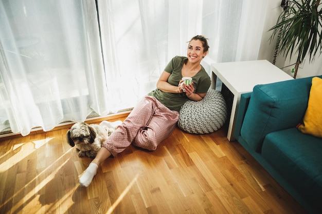 Junge lächelnde Frau, die am gemütlichen Morgen zu Hause mit ihrem Shih Tzu-Hund Kaffee genießt.