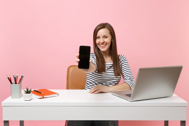 Junge lächelnde Frau, die auf Kamerahandy mit leerem leerem Bildschirmarbeit am weißen Schreibtisch mit zeitgenössischem PC-Laptop einzeln auf pastellrosa Hintergrund zeigt. Erfolg Geschäftskarriere. Platz kopieren.