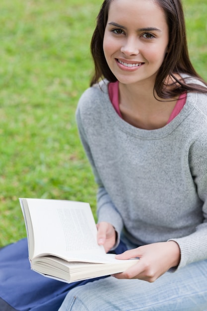 Junge lächelnde Frau, die ein Buch in einem Park beim Betrachten der Kamera liest