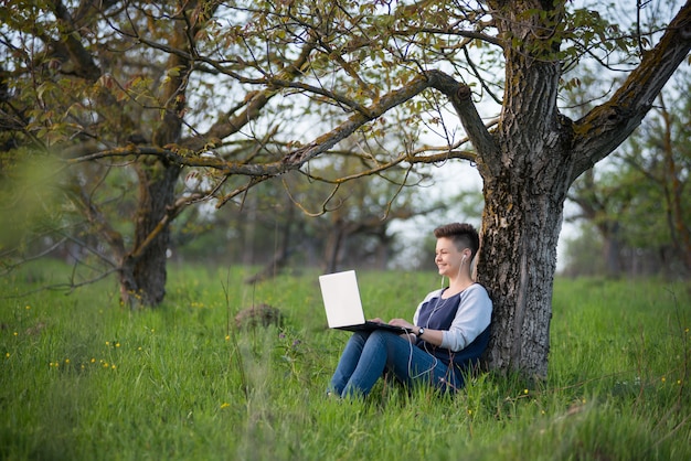 Junge lächelnde Frau, die unter dem Baum im Park mit ihrem Laptop sitzt