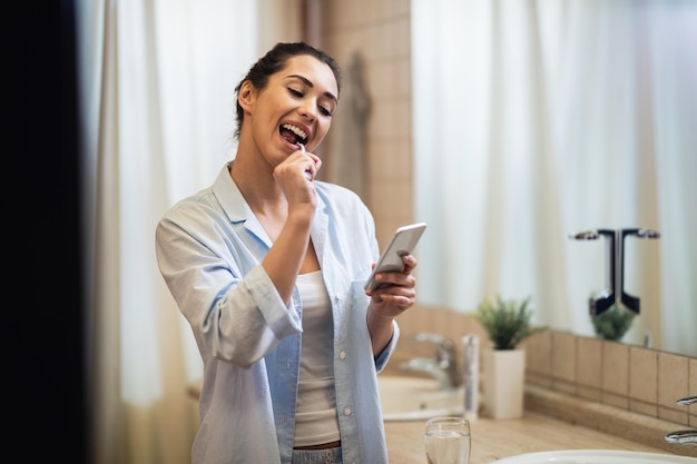 Junge lächelnde Frau, die Zähne putzt und SMS auf dem Handy im Badezimmer liest