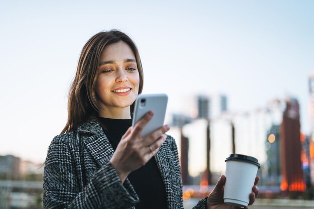 Junge lächelnde Frau im Mantel mit Kaffeetasse mit Handy in der abendlichen Stadtstraße