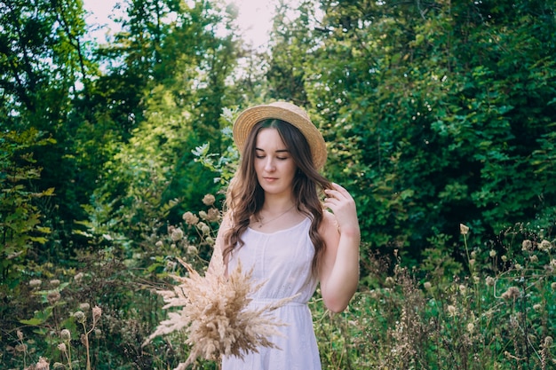 Junge lächelnde Frau in einem Strohhut auf einem Hintergrund des grünen Sommerwaldes. Schönes Mädchen mit einem Strauß getrockneter Blumen auf einem Spaziergang. Mädchen in einem Sommerkleid.