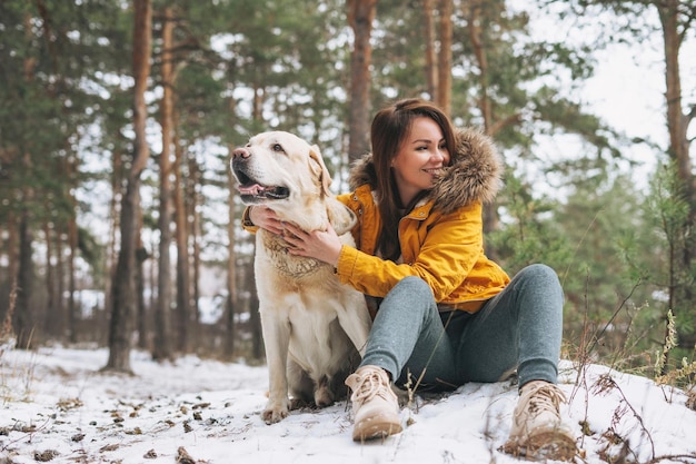 Junge lächelnde Frau in gelber Jacke mit großem freundlichen weißen Hund Labrador, der im Winterwald spaziert