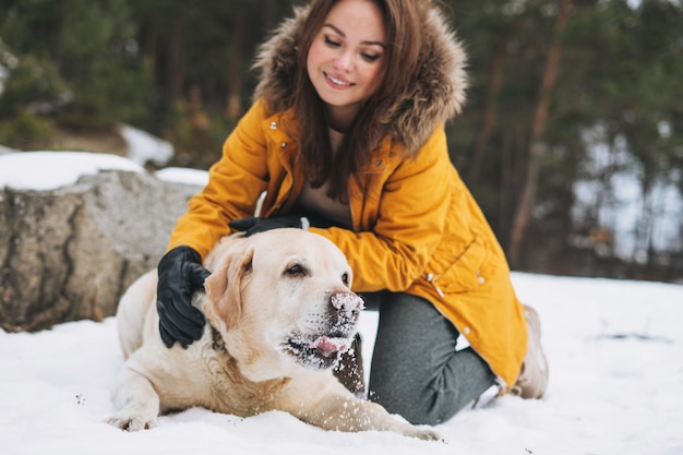 Junge lächelnde Frau in gelber Jacke mit großem freundlichen weißen Hund Labrador, der im Winterwald spaziert