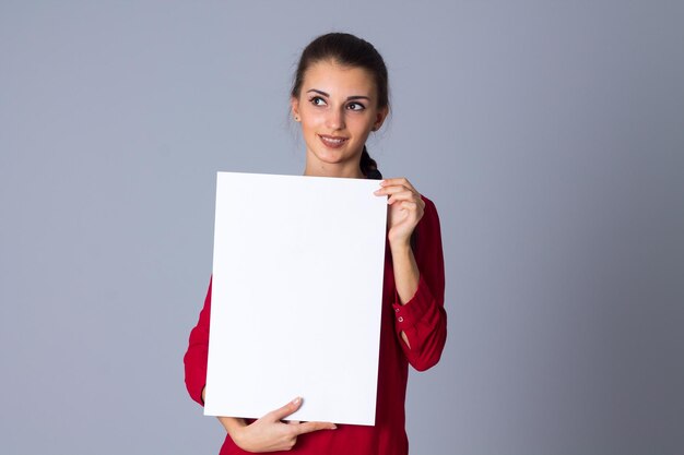 Junge lächelnde Frau in roter Bluse mit Zopf, die weißes Blatt Papier im Studio zeigt