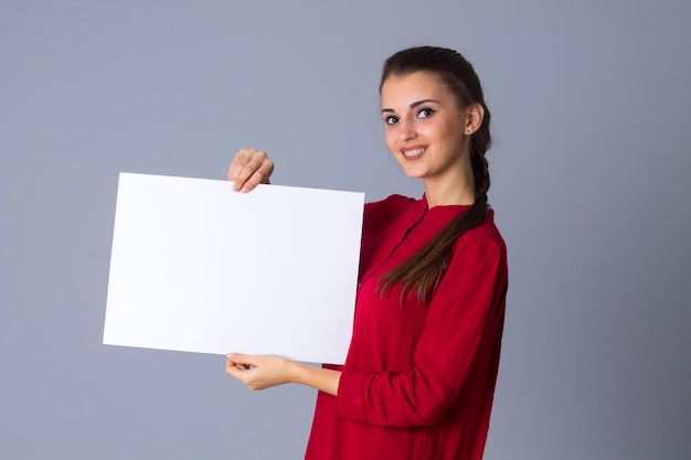 Junge lächelnde Frau in roter Bluse mit Zopf, die weißes Blatt Papier im Studio zeigt