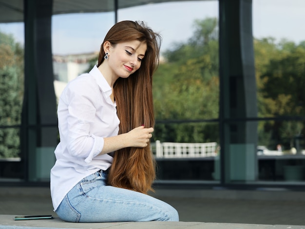 junge lächelnde frau in weißer bluse und jeans berührt ihr langes haar mit der hand