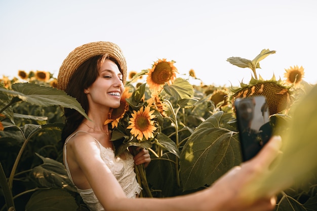 Junge lächelnde Frau mit Hut macht Selfie-Foto im Sonnenblumenfeld