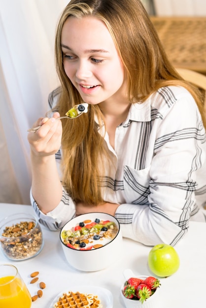 Junge lächelnde Frau mit leckerem gesundem Müsli mit Obst und Joghurt zum Frühstück zu Hause