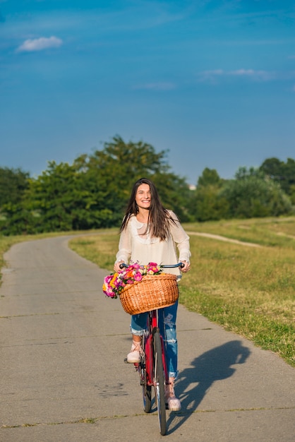 Junge lächelnde Frau reitet ein Fahrrad mit einem Korb voller Blumen in der Landschaft