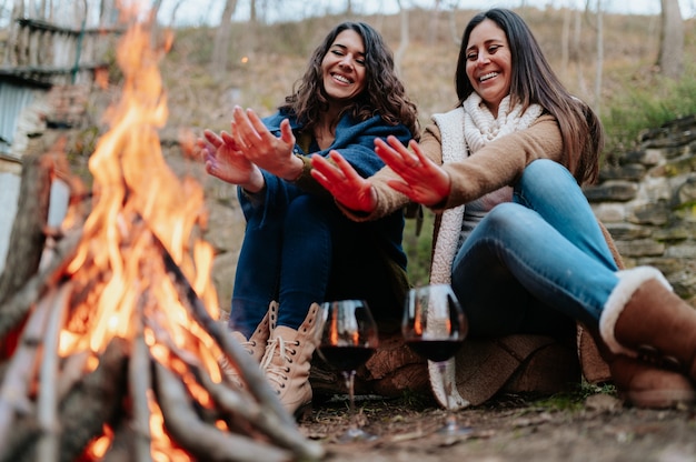 Junge lächelnde Frauen, die sich neben dem Feuer erwärmen. Gläser Rotwein. Lagerfeuer, Aktivitäten im Freien, Entspannung, Zusammengehörigkeitskonzept.