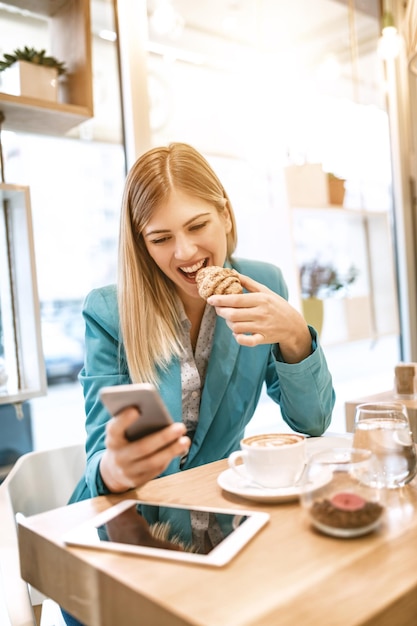 Junge lächelnde Geschäftsfrau auf einer Pause in einem Café. Er frühstückt und benutzt Smartphone.