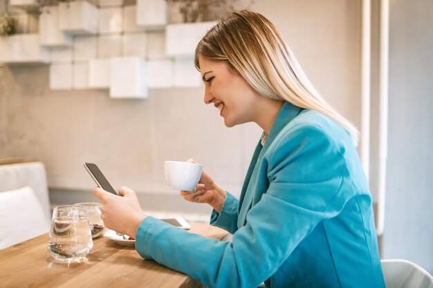 Junge lächelnde Geschäftsfrau auf einer Pause in einem Café. Er trinkt Kaffee und benutzt Smartphone.