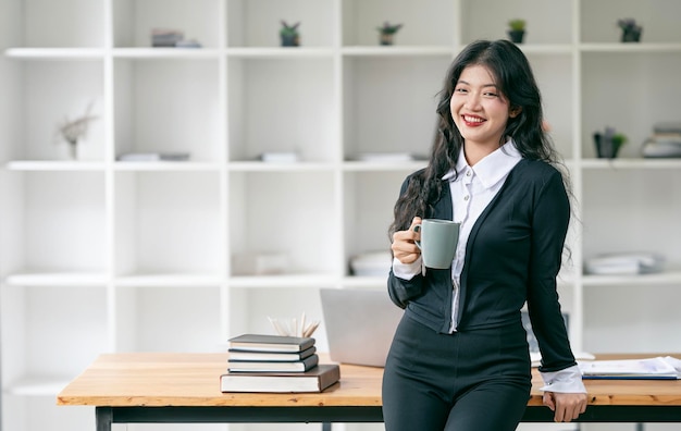 Junge lächelnde Geschäftsfrau hält einen Becher in der Hand und steht an ihrem Schreibtisch im Büro und blickt in die Kamera