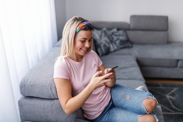 Junge lächelnde herrliche Hausfrau, die auf Couch im Wohnzimmer sitzt, eine Pause macht und Smartphone benutzt.