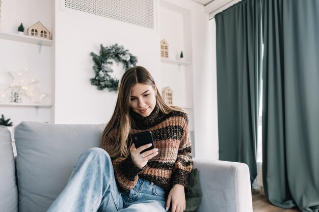 Junge lächelnde kaukasische Frau, die Handy in den Ferien zu Hause auf dem Sofa verwendet.