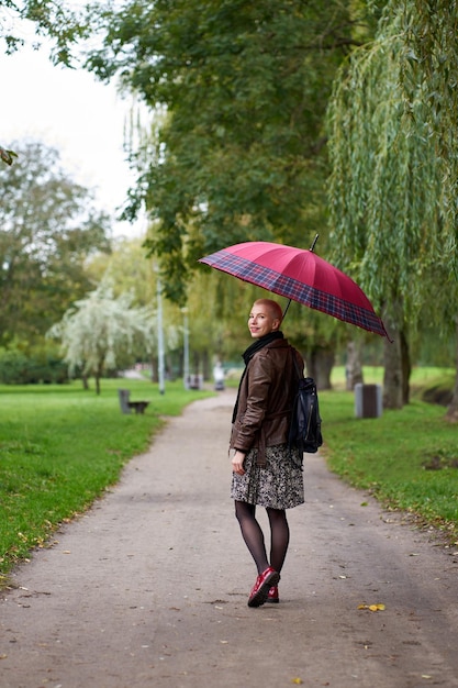 Foto junge lächelnde kurzhaarige blonde frau geht im herbstpark mit rotem regenschirm blick von hinten