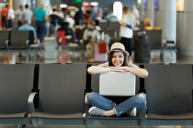 Junge lächelnde reisende Touristenfrau, die mit Laptop mit gekreuzten Beinen sitzt, während sie in der Lobbyhalle am internationalen Flughafen wartet?