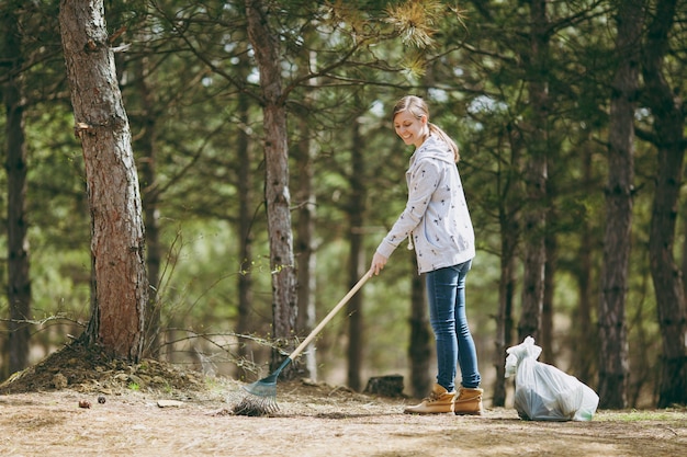 Junge lächelnde schöne Frau, die Rechen für die Müllabfuhr in der Nähe von Müllsäcken im Park oder Wald säubert und verwendet. Problem der Umweltverschmutzung. Stoppen Sie Naturmüll, Umweltschutzkonzept.