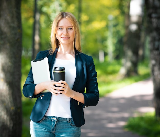 Junge lächelnde schöne Frau im Park mit Tablet-Gadget und drahtlosen Kopfhörern, die Fernarbeit mit grünem Energiekonzept genießen