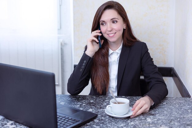 Junge lächelnde schöne Geschäftsfrau, die im Büro sitzt und am Laptop arbeitet und an einem Telefon spricht