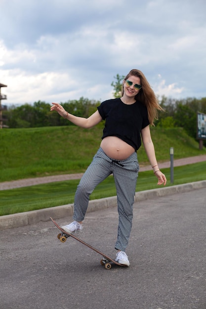 Junge lächelnde schwangere Frau in schwarzem T-Shirt und herzförmiger Sonnenbrille beim Skateboarden auf der Straße auf dem Hintergrund des grünen Rasens