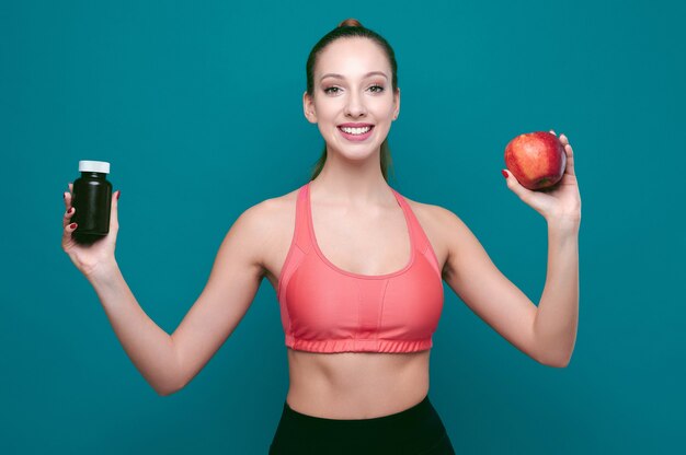 Junge lächelnde sportliche Frau zeigt Apfel und Pillen oder Drogen auf grünem isoliertem Hintergrund