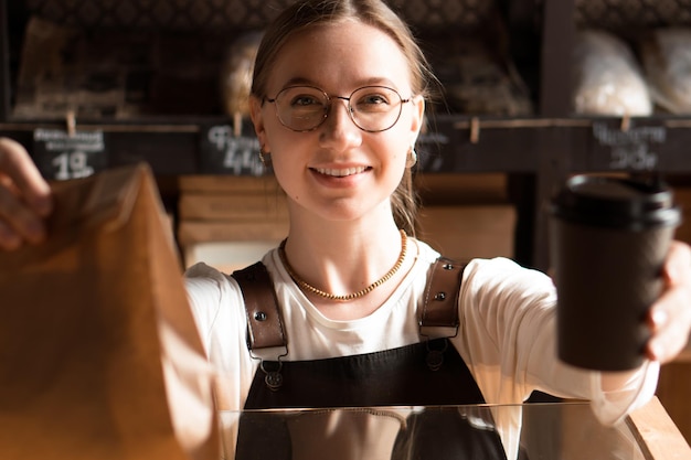 Junge lächelnde Verkäuferin in der Bäckerei mit Papiertüte und Kaffeetasse zum Mitnehmen im Café