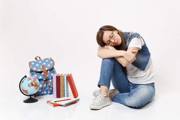 Junge, lässige, müde, entspannte Studentin in Denim-Kleidung, die in der Nähe von Globus, Rucksack, Schulbüchern sitzt, isoliert