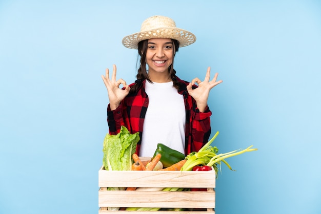 Junge Landwirtin, die frisches Gemüse in einem hölzernen Korb hält, der ein OK-Zeichen mit den Fingern zeigt