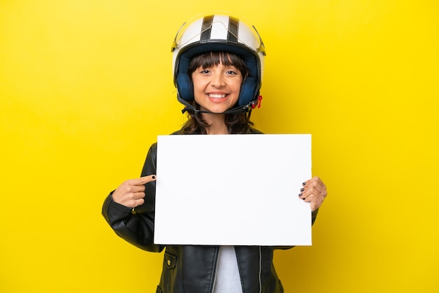 Junge lateinamerikanische Frau mit Motorradhelm isoliert auf gelbem Hintergrund, die ein leeres Plakat mit fröhlichem Gesichtsausdruck hält und darauf zeigt