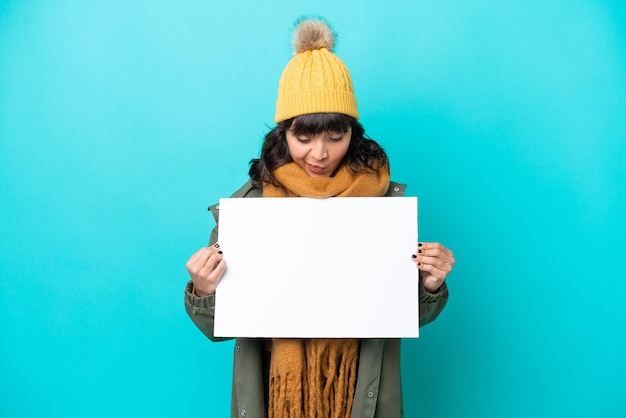 Junge lateinamerikanische Frau mit Winterjacke isoliert auf blauem Hintergrund, die ein leeres Plakat hält und es ansieht