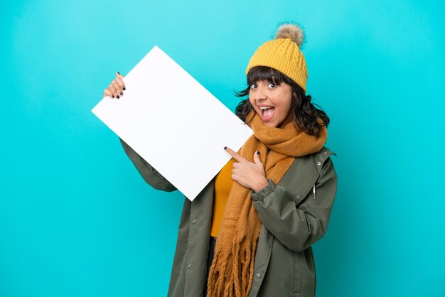 Junge lateinamerikanische Frau mit Winterjacke isoliert auf blauem Hintergrund, die ein leeres Plakat mit fröhlichem Gesichtsausdruck hält und darauf zeigt