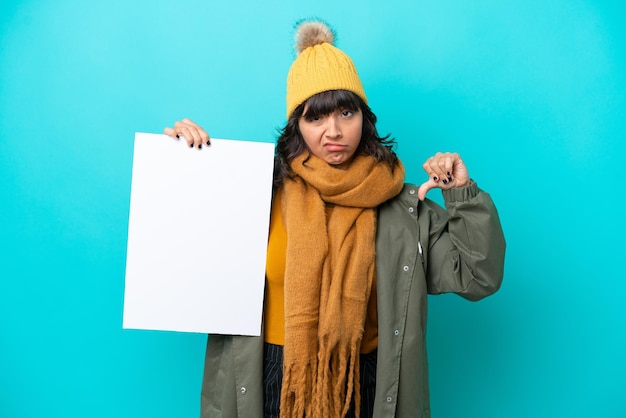 Junge lateinamerikanische Frau mit Winterjacke isoliert auf blauem Hintergrund mit leerem Plakat und schlechtem Signal