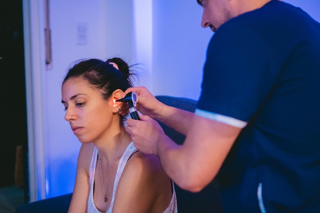 Junge lateinamerikanische Frau zu Hause lässt ihre Ohren von einem Besuchsarzt mit einem Otoskop untersuchen