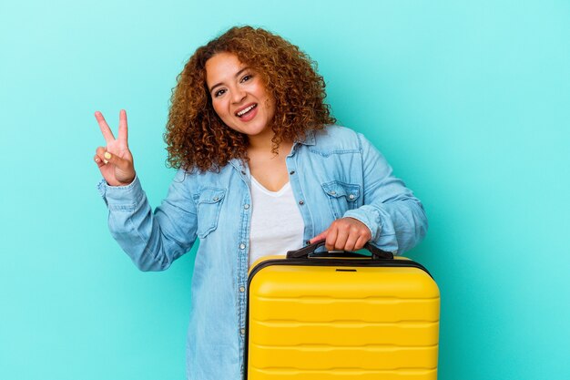 Junge lateinamerikanische Reisende kurvige Frau, die einen Koffer isoliert auf blauem Hintergrund hält, fröhlich und sorglos, der ein Friedenssymbol mit den Fingern zeigt.