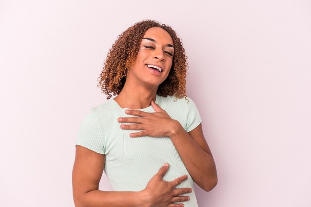 Junge lateinamerikanische transsexuelle Frau, die auf rosafarbenem Hintergrund isoliert ist, lacht glücklich und hat Spaß, die Hände auf dem Bauch zu halten.