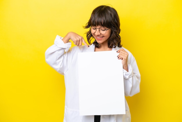 Junge lateinische Ärztin isoliert auf gelbem Hintergrund, die ein leeres Plakat mit glücklichem Ausdruck hält und darauf zeigt