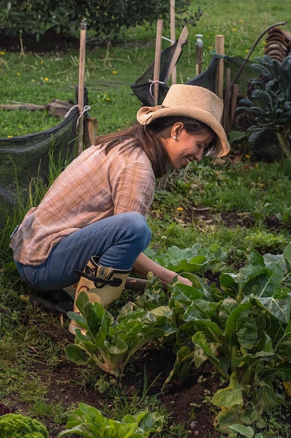 Junge lateinische Bäuerin mit Hut und Denim hockt und reinigt den Gartenboden, umgeben von grünem Gemüse