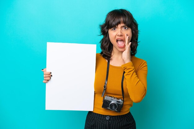 Junge lateinische Fotografin isoliert auf blauem Hintergrund, die ein leeres Plakat hält und schreit