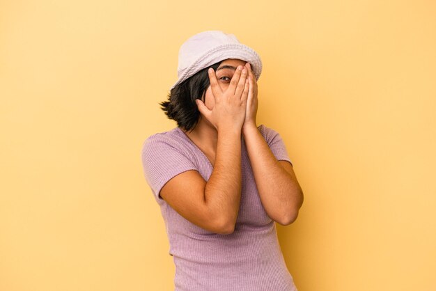 Junge lateinische Frau, die auf gelbem Hintergrund isoliert ist, blinzelt erschrocken und nervös durch die Finger.